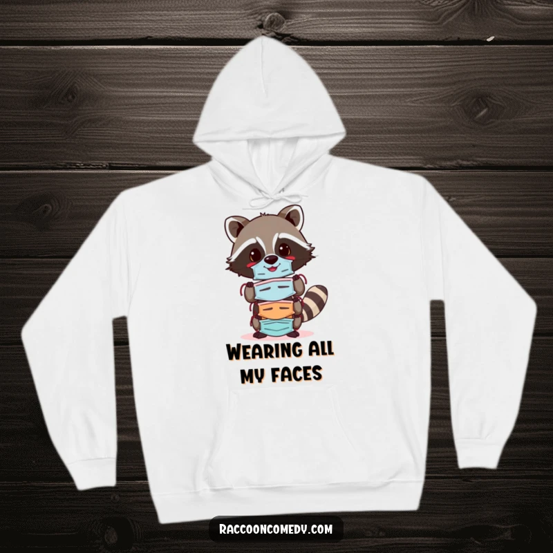Funny raccoon hoodie showcasing a cheerful raccoon balancing a stack of colorful masks, perfect for cozying up with a playful attitude.